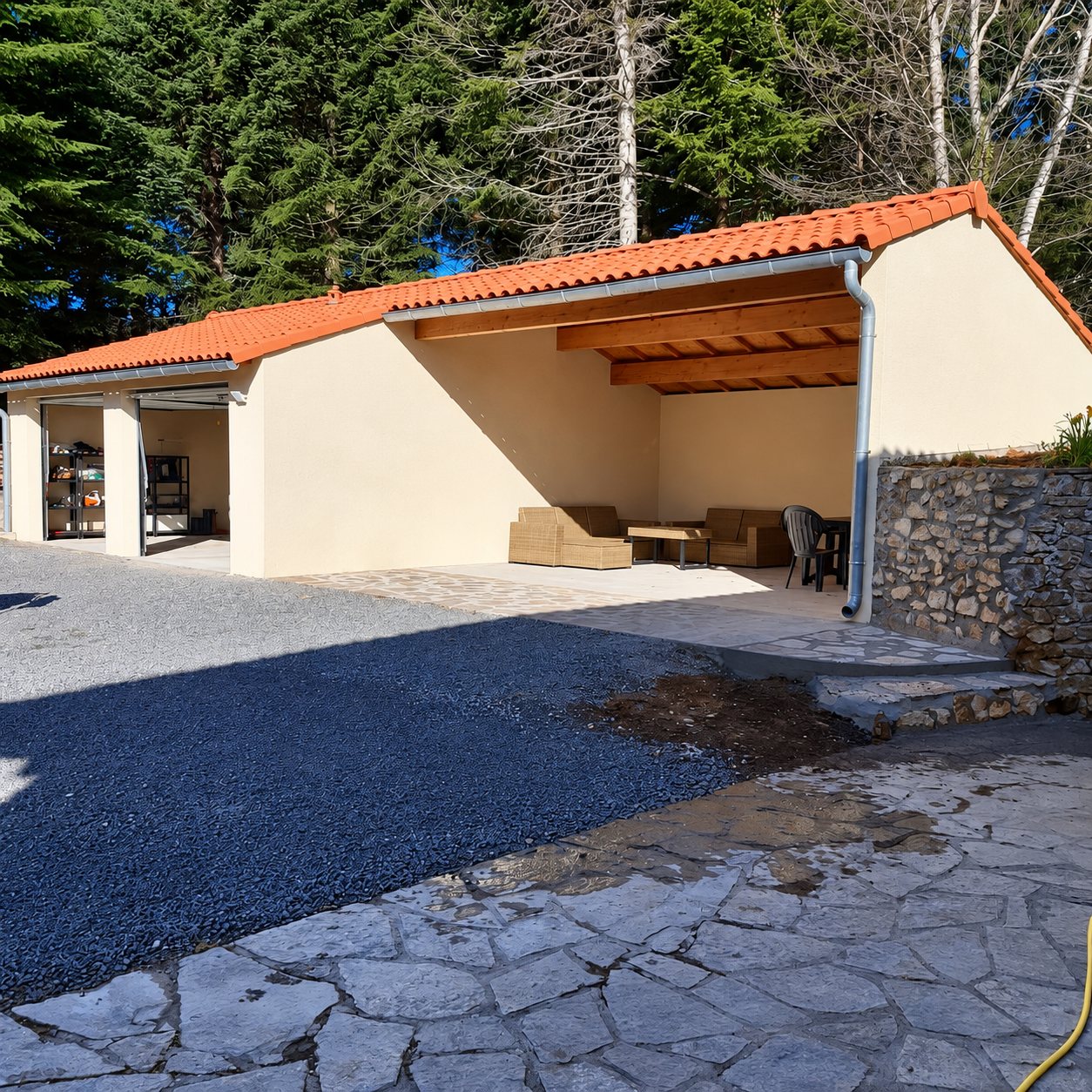 Un bâtiment récent aux murs crème et à la toiture en tuiles oranges, avec une partie ouverte aménagée en salon de jardin, entouré de gravier, de dalles de pierre et de grands arbres.
