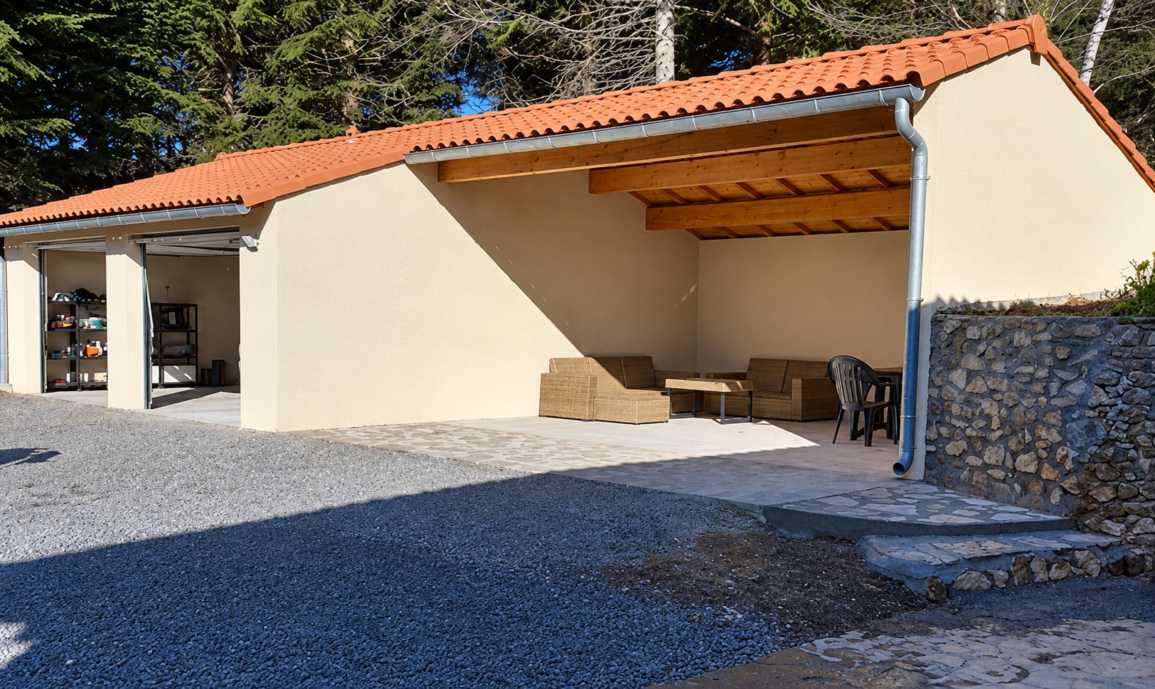 Bâtiment aux murs crépis beige et à la toiture de tuiles orange, avec une terrasse couverte meublée d'un salon de jardin en rotin, un muret de pierre et une allée de gravier.