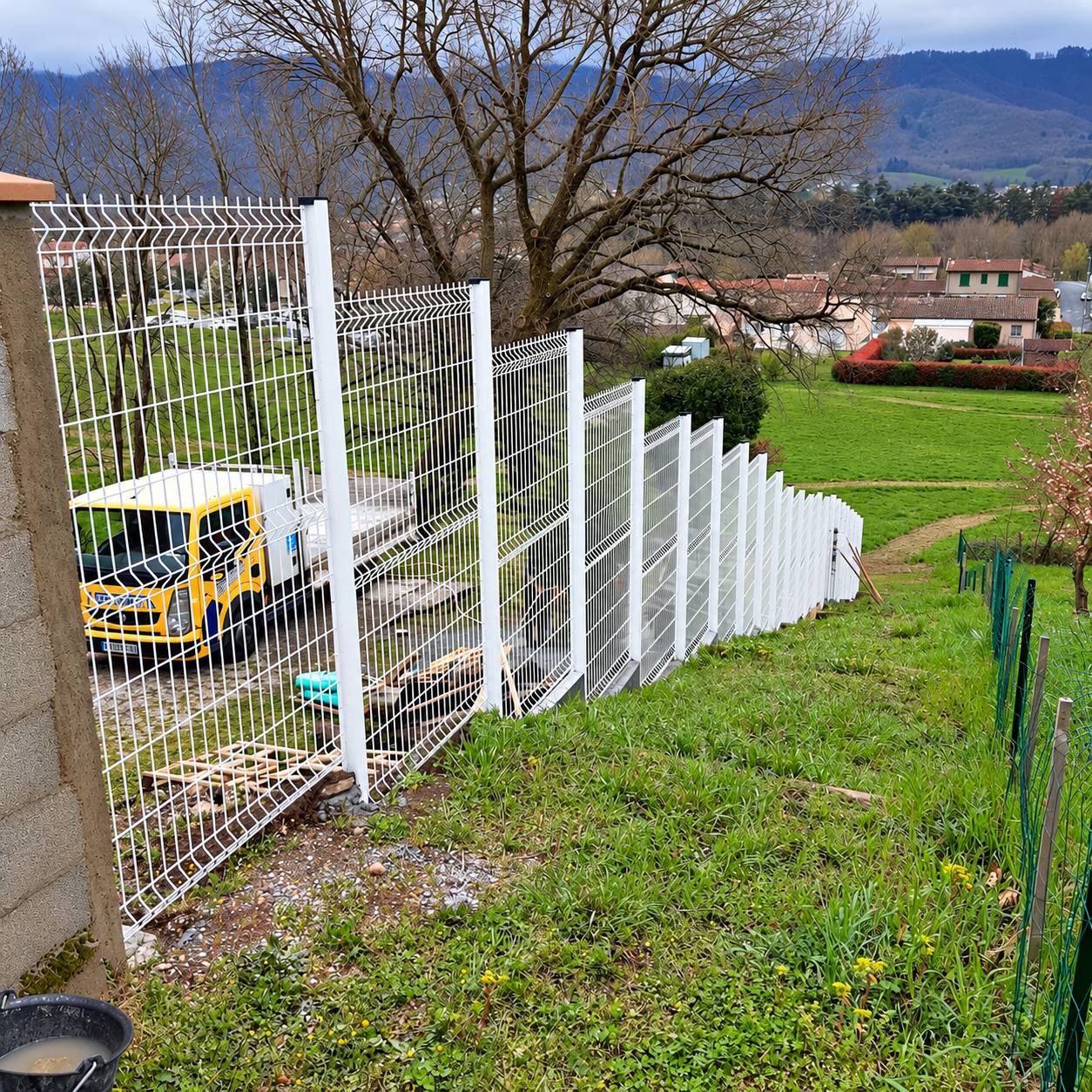 Une clôture en panneaux de grillage rigide blanc longe un talus herbeux, avec un camion jaune garé derrière et des maisons dans la vallée.