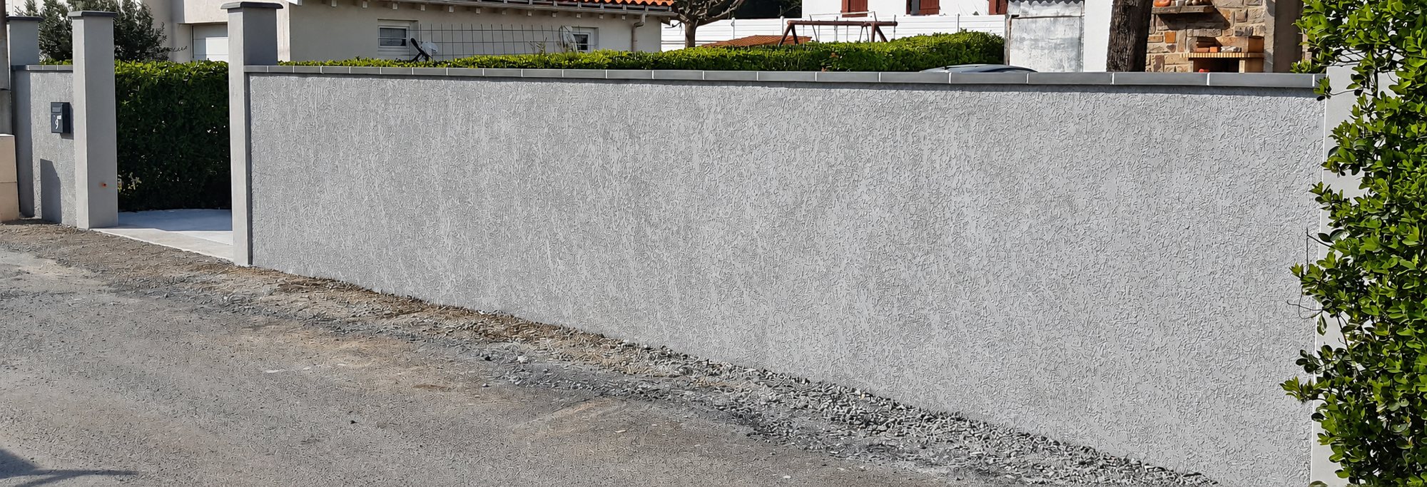 Mur de clôture en crépi gris avec piliers et portail le long d’une rue