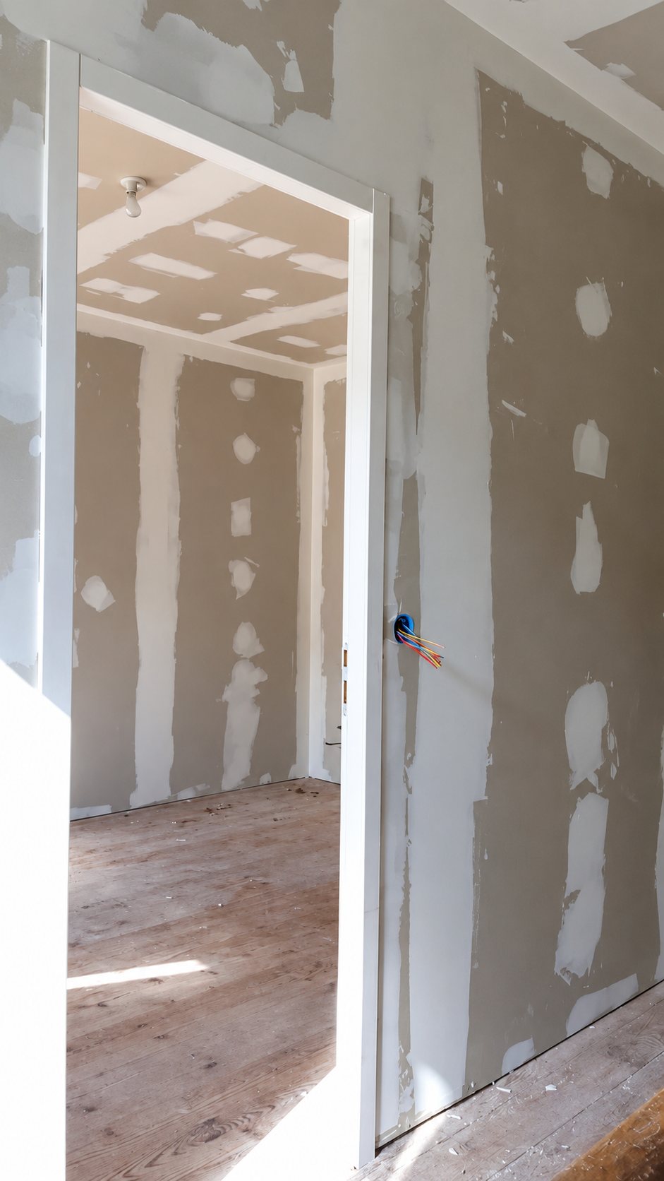 Vue à travers un cadre de porte blanc sur une pièce en travaux dont les murs et le plafond en plaques de plâtre sont en cours de jointoiement, avec des fils électriques apparents.
