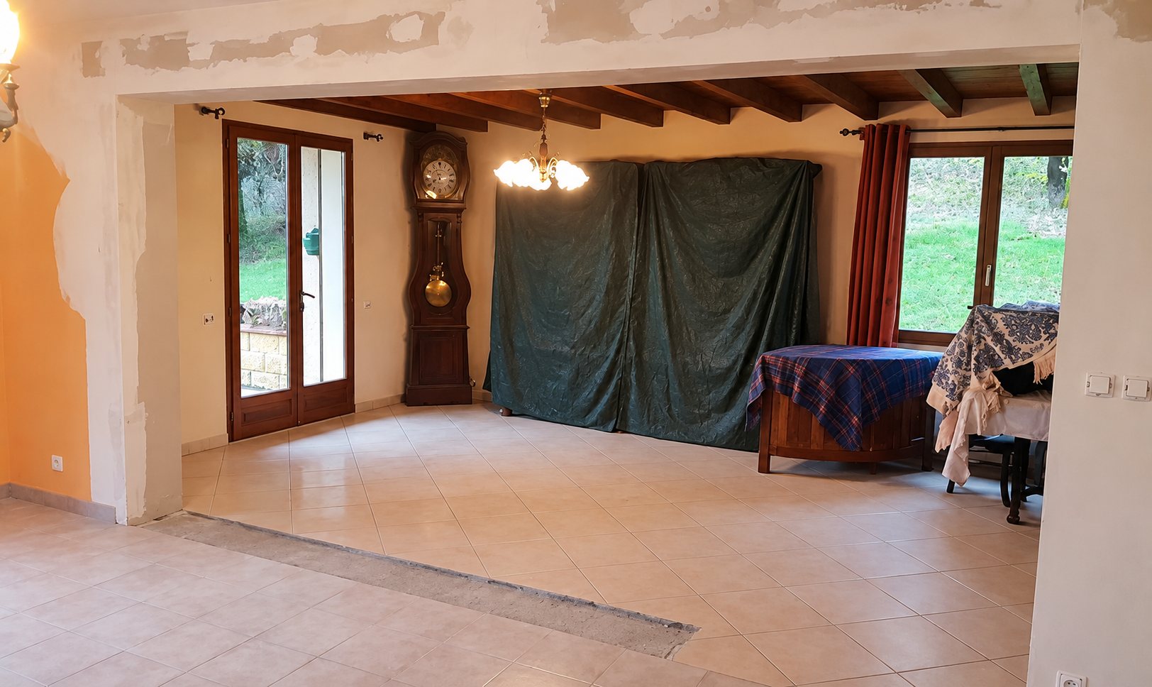 Une grande pièce à vivre en cours de rénovation, avec murs en plâtre brut, poutres apparentes, sol carrelé clair, horloge comtoise et grandes fenêtres donnant sur un jardin.