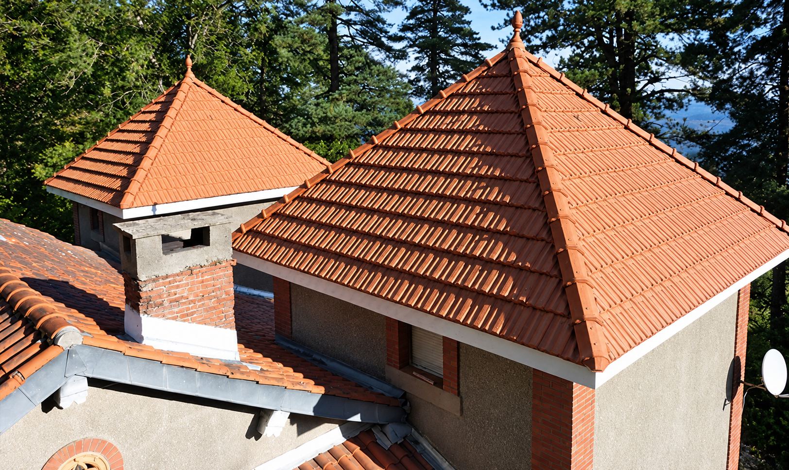 Vue en hauteur d'une toiture à pans en tuiles canal rouges fraîchement posées sur une maison entourée de pins.